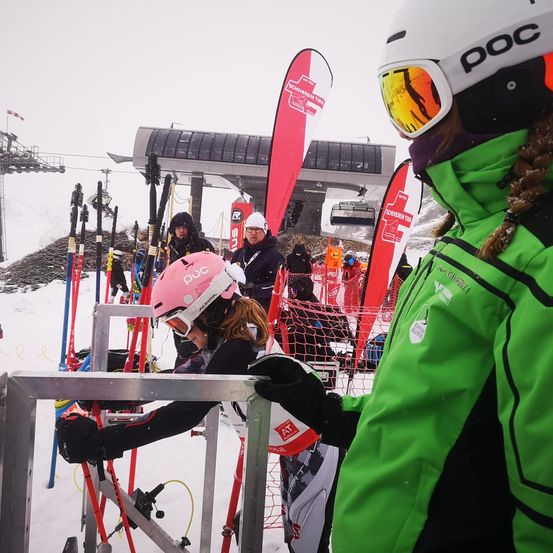 Eine Frau mit einem rosa Helm bereitet sich zum Skifahren vor und steht in einer Warteschlange an einem Skilift. Dahinter sitzt ein weiterer Skifahrer auf einem Stuhl, und mehrere andere warten. Rote Banner sind im Hintergrund zu sehen, und ein Gebäude mit einem Glasdach ist sichtbar.