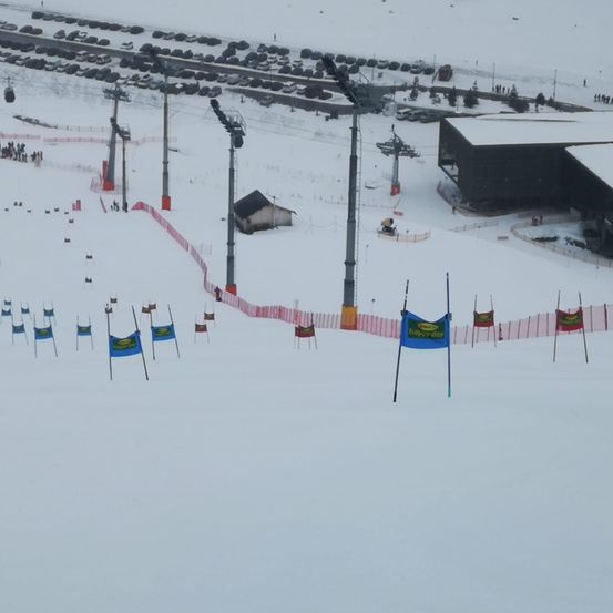 Eine verschneite Skipiste mit Fahnen, die den Kurs markieren. Ein Gebäude und ein Parkplatz sind sichtbar. Mehrere Skilifte befinden sich im Hintergrund.