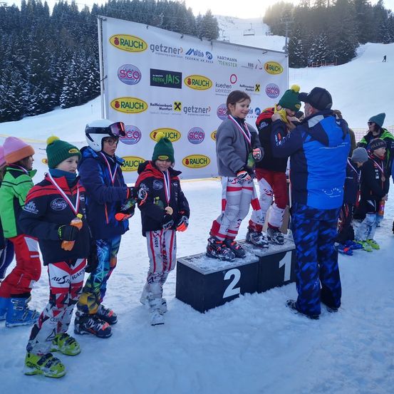 Eine Gruppe von Kindern steht auf einem Podest bei einer Skiveranstaltung. Sie tragen Winterkleidung und Skischuhe. Das Podest hat die Zahlen 1, 2 und 3. Dahinter befindet sich ein Banner mit Logos und Text.