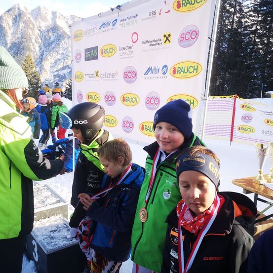 Vier junge Jungen in Winterkleidung und mit Medaillen stehen neben einem Banner mit verschiedenen Logos, wahrscheinlich in einem verschneiten Gebirgsgebiet. Dahinter überreicht eine Person Auszeichnungen.