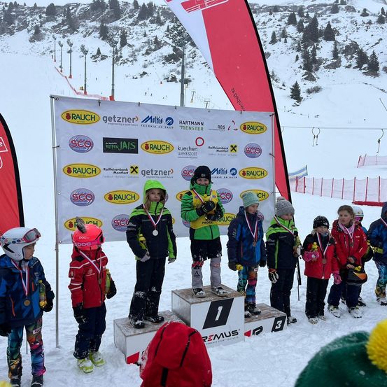 Kinder stehen auf einem Podest in einer verschneiten Landschaft, tragen Winterkleidung und halten Skistöcke. Ein großes Banner hinter ihnen zeigt verschiedene Logos und Text.