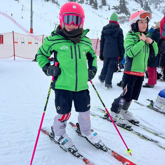 Zwei Kinder in grünen Skijacken und Helmen stehen auf Skiern im Schnee und halten Skistöcke. Sie befinden sich auf einer Skipiste mit Netzzaun und Menschen im Hintergrund.