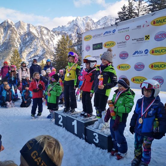 Kinder in Skiausrüstung stehen auf einem Podest in einem verschneiten Bereich, mit Bergen im Hintergrund. Einige halten Flaschen und haben Medaillen. Eine Wand mit Werbung steht hinter ihnen.