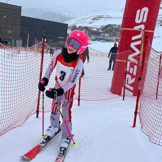 Eine junge Skifahrerin mit rotem Helm und Schneebrille steht auf schneebedecktem Boden mit Skistöcken. Hinter ihr befindet sich ein roter Banner mit dem Wort 'SUI'. Im Hintergrund sind ein Gebäude und verschneite Berge zu sehen.