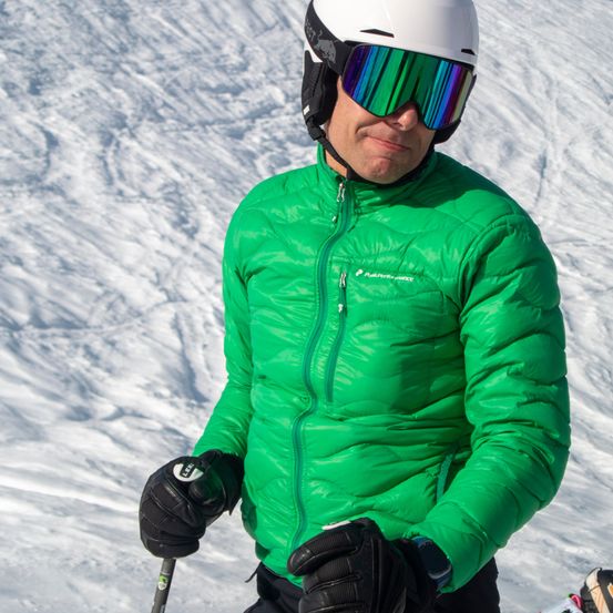 Ein Skifahrer in einer grünen Jacke, weißem Helm und mit Brille steht auf einem verschneiten Hang und hält Skistöcke.