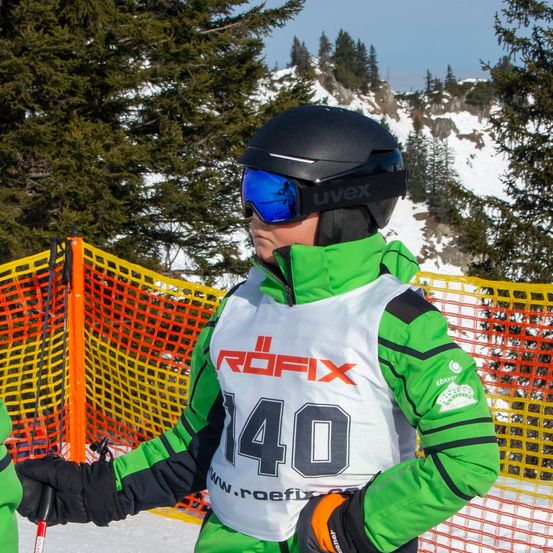 Ein Skifahrer in einem grünen und schwarzen Outfit mit der Nummer 140 steht vor einem gelben Zaun auf einer verschneiten Piste.