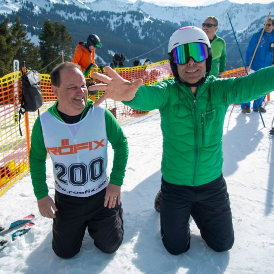 Zwei Männer knien im Schnee mit Winterausrüstung, einer trägt einen grünen Mantel mit ROFIX 200, der andere einen Helm und eine Schutzbrille. Sie lächeln und haben Skier hinter sich. Schneebedeckte Berge im Hintergrund.