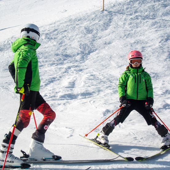 Zwei Skifahrer sind auf einem verschneiten Hang, einer trägt eine grüne Jacke und der andere einen pinken Helm. Beide halten Skistöcke und scheinen das Skifahren zu lernen.