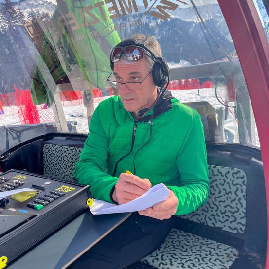Ein Mann in einer grünen Jacke sitzt in einer Kabine, trägt Kopfhörer, hält ein Papier in der Hand und schaut auf ein Bedienfeld. Hinter ihm sind Berge und Schnee zu sehen.