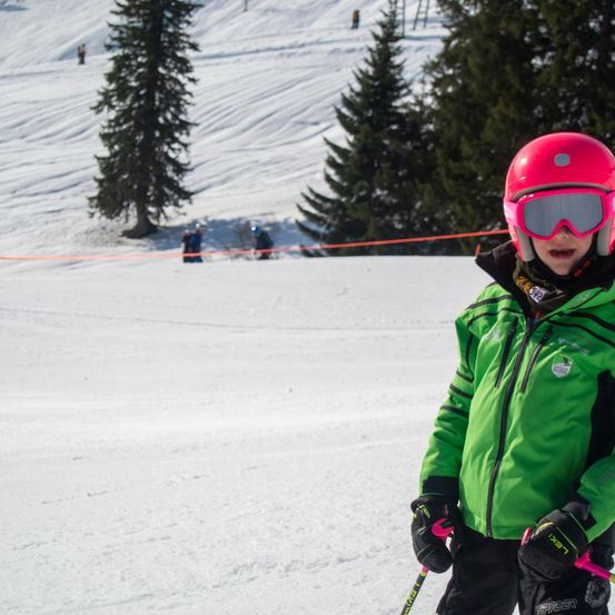 Ein junger Skifahrer in grüner und rosa Skiausrüstung steht auf einem verschneiten Hang mit immergrünen Bäumen im Hintergrund.