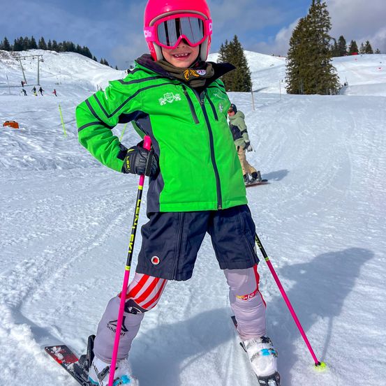 Ein junges Kind steht auf einem verschneiten Hang, trägt eine grüne Skijacke und eine pinke Schutzbrille. Es hält Skistöcke und schaut nach vorne. Im Hintergrund sind andere Skifahrer auf dem Hang, und Bäume säumen den Horizont.