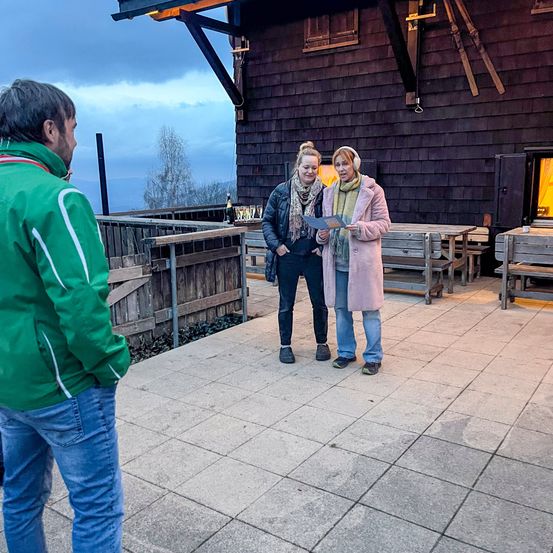 Zwei Frauen und ein Mann stehen auf einer gefliesten Terrasse. Die Frauen schauen auf etwas, das der Mann in der Hand hält, während der Mann sie ansieht. Hinter ihnen steht eine Holzkonstruktion mit Tisch und Stühlen.