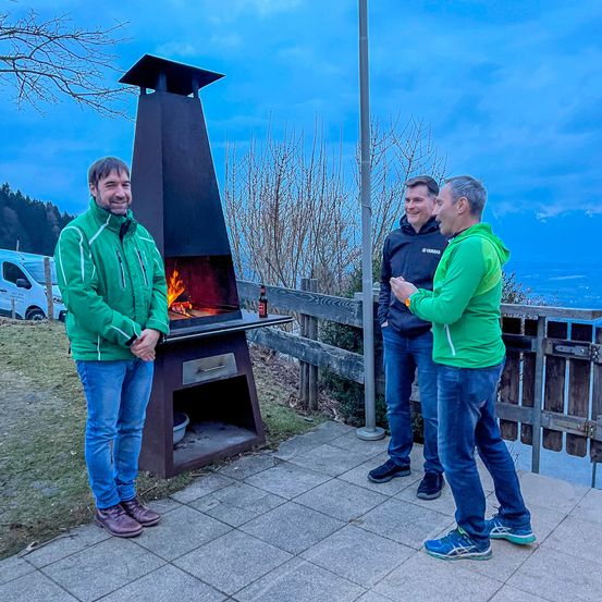 Drei Männer stehen neben einem schwarzen Außenkamin mit Feuer im Inneren. Sie sind in grünen Jacken und Jeans gekleidet. Hinter ihnen steht ein Holzzaun und ein weißer Lieferwagen.