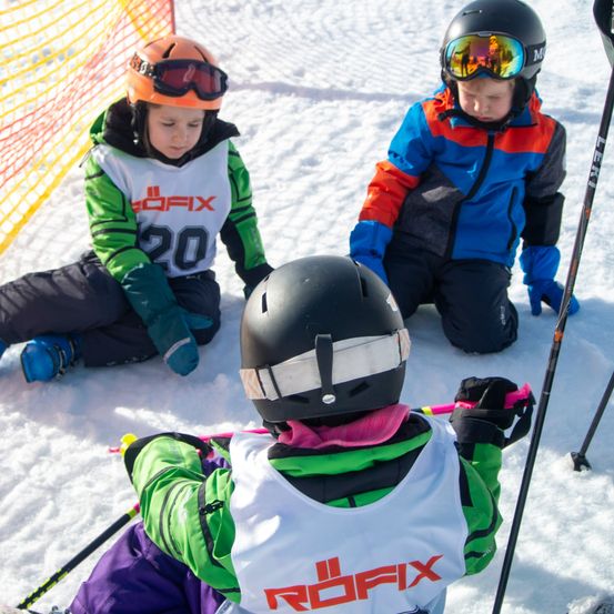 Drei Kinder sitzen im Schnee und tragen Skiausrüstung und Helme. Eines trägt eine grün-weiße 'ROFIX'-Jacke, ein anderes eine blau-rote Jacke und das dritte einen schwarzen Helm. Skistäbe sind in der Nähe.