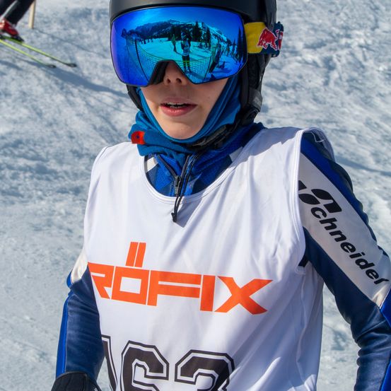 Eine Person mit Helm, Schutzbrille und blauem Schal steht auf einem verschneiten Hang. Die Person trägt ein weißes Trikot mit orangefarbenen Buchstaben und der Nummer 162.