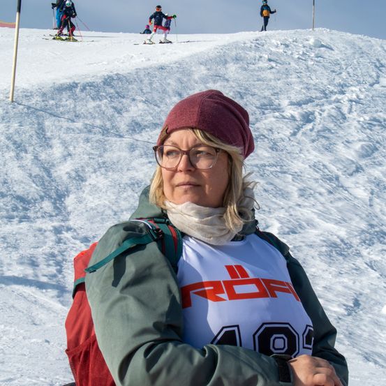 Eine Frau mit Brille und roter Mütze sitzt auf einem verschneiten Hügel. Hinter ihr steigen drei Skifahrer den Hang hinunter.