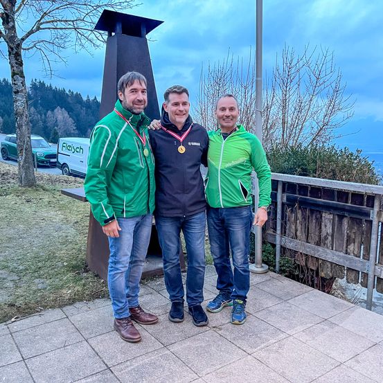 Drei Männer stehen auf einer Terrasse, tragen grüne Jacken und Medaillen, mit einem Turm und einem Blick auf Berge im Hintergrund.