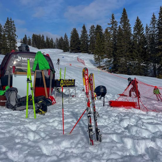 Ein Ski-Equipment-Setup auf schneebedecktem Gelände mit Bäumen im Hintergrund, eine Digitaluhr mit der Anzeige 00:00 und Personen, die einen Hang hinunterfahren.