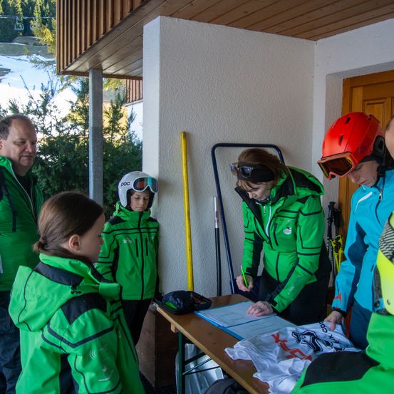 Eine Gruppe von Menschen in grünen Skijacken versammelt sich um einen Tisch mit Papieren in der Nähe eines verschneiten Berges. Eine Frau schreibt auf ein Stück Papier.