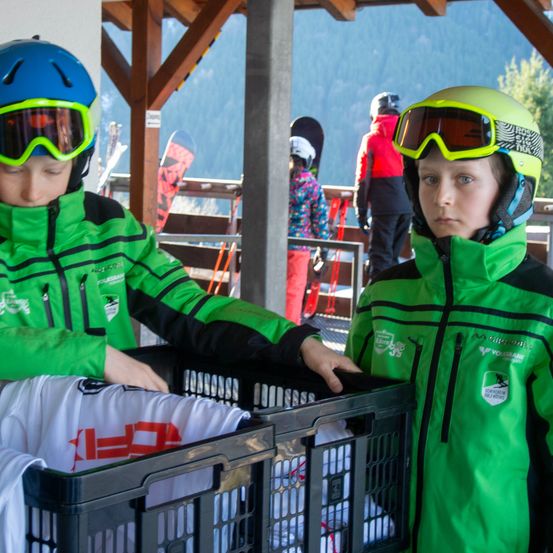 Zwei Kinder in grünen Skijacken und Helmen stehen neben einem Korb mit weißen T-Shirts. Sie befinden sich in einem verschneiten Außenbereich mit Holzkonstruktionen und Bergen im Hintergrund.