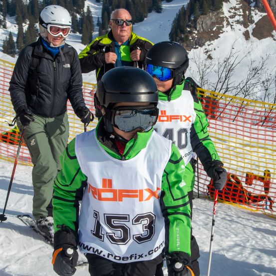 Ein Skifahrer mit der Nummer 153, der eine grün-weiße Jacke trägt, steht auf einer verschneiten Piste. Zwei andere Personen, die Helme und Schutzbrillen tragen, stehen hinter ihm. Einer von ihnen hält einen Skistock. Ein Netzzaun befindet sich hinter ihnen.