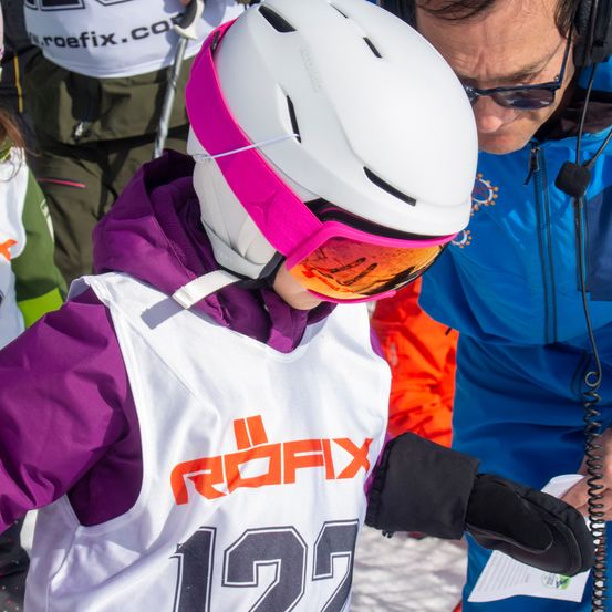 Ein Skifahrer in einem weißen Helm mit der Nummer 125 auf dem Jackett wird an einem verschneiten Tag von einem Trainer überprüft.