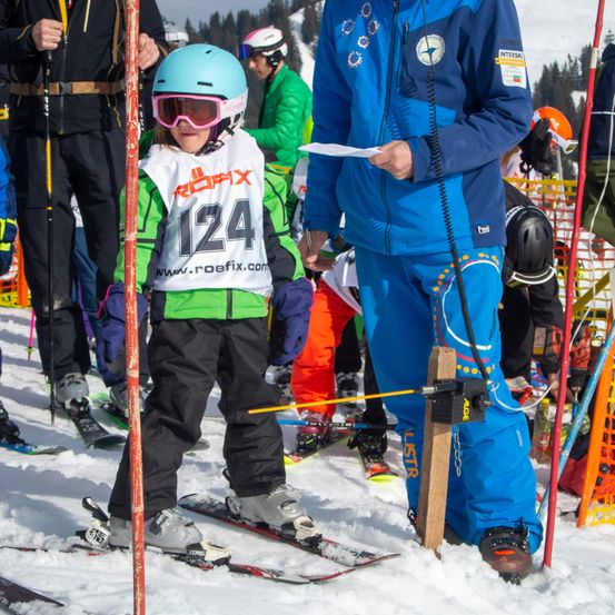 Ein Kind mit der Nummer 124 steht auf Skiern, ein Trainer hält in der Nähe ein Blatt Papier. Sie befinden sich in einem verschneiten Bereich, umgeben von anderen Skifahrern.
