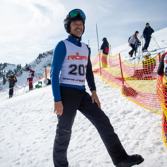 Ein Mann mit einem Nummer-20-Trikot und Helm steht auf einer verschneiten Piste. Andere Skifahrer befinden sich hinter einer Netzbarriere im Hintergrund.