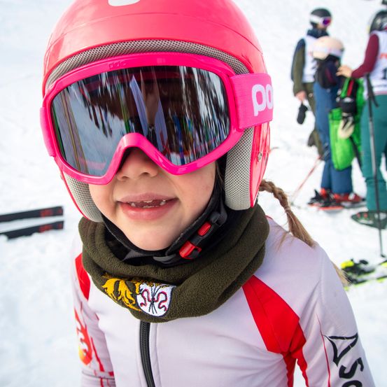 Ein Kind mit einem rosa Helm und Schutzbrille lächelt an einem verschneiten Tag. Es trägt einen weißen und roten Skianzug. Im Hintergrund sind andere Skifahrer zu sehen.