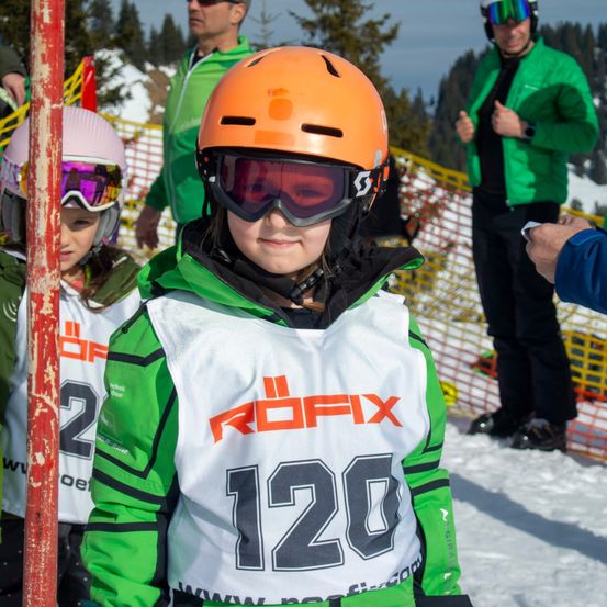 Ein junges Kind in einem grün-weißen Jacke mit der Nummer 120 steht auf schneebedecktem Boden. In der Nähe sind Erwachsene in grünen und schwarzen Jacken anwesend.