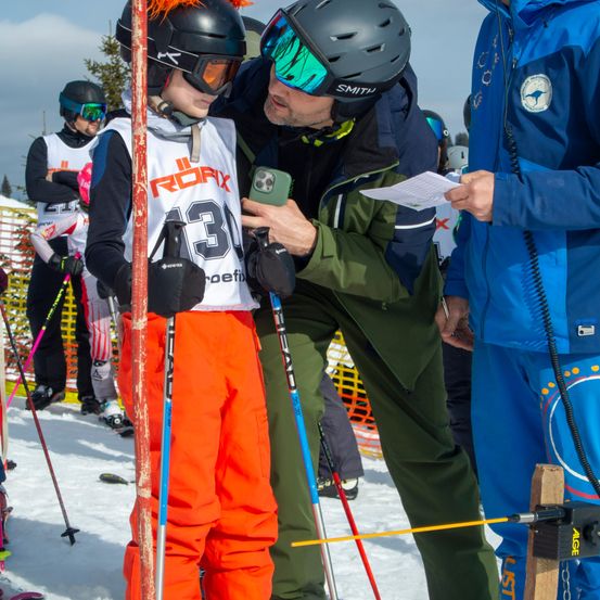 Ein Kind in einer orangefarbenen Kleidung mit der Nummer 33 steht mit Skiern, während ein Erwachsener mit Helm und Brille ihm hilft, ein Handy zu halten. In der Nähe hält ein anderer Erwachsener ein Stück Papier. Sie befinden sich in einem verschneiten Bereich mit anderen Skifahrern.