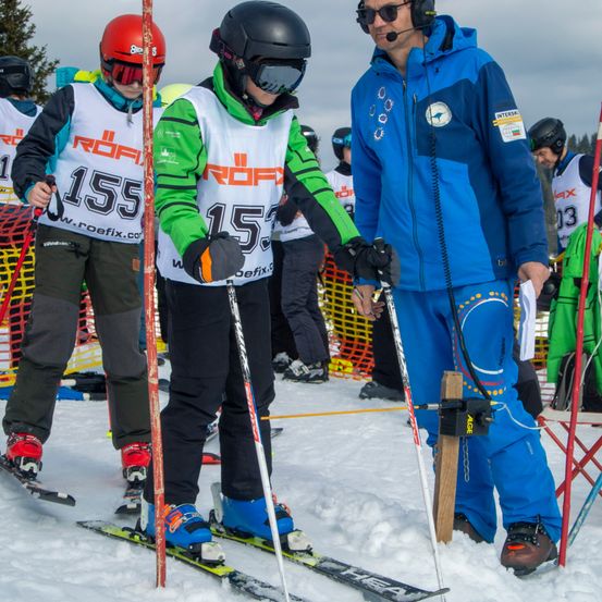 Skifahrer mit Helmen und Schutzbrillen bereiten sich auf ein Rennen auf einem verschneiten Feld vor. Ein Mann hält einen Stock und Papier.