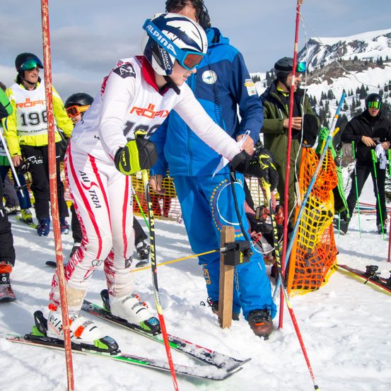 Ein Skifahrer in einem weißen Skianzug bereitet sich auf einen Abfahrtslauf auf einem verschneiten Berg vor. Ihn umgeben mehrere Personen in farbenfrohen Skianzügen.
