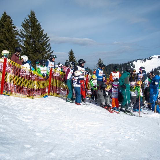 Eine Gruppe von Skifahrern und Snowboardern versammelt sich auf einem verschneiten Hang, mit Helmen und Skianzügen bekleidet. Einige tragen nummerierte Tags, und ein gelber Zaun steht hinter ihnen. Bäume sind im Hintergrund.