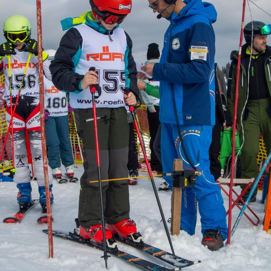 Skifahrer mit nummerierten Trikots stehen im Schnee, einige halten Skistöcke. Ein Mann in einem blauen und ein anderer in einem grünen Jackett stehen in der Nähe von ihnen, der Mann in Blau hält ein Stück Papier.