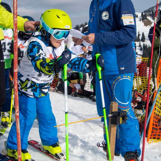 Ein junger Skifahrer, der einen Helm und eine Brille trägt, erhält von einem Trainer auf einem verschneiten Hang ein Zertifikat. Im Hintergrund befinden sich andere Skifahrer, Berge und ein Zaun.