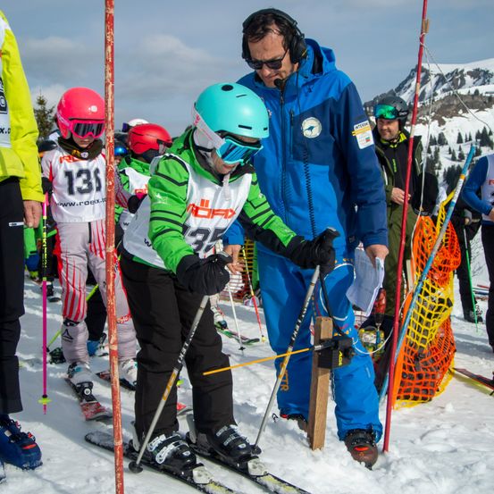 Eine Gruppe junger Skifahrer, die Helme und Schutzbrillen tragen, übt das Skifahren auf einer verschneiten Piste. Ein Lehrer hält das Skistöcken eines Kindes. Im Hintergrund befindet sich ein Berg und orange Netze.