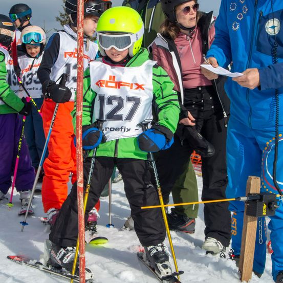 Eine Gruppe junger Skifahrer, in grünen und weißen Uniformen, steht auf schneebedecktem Boden. Ein Kind, Nummer 127, hält Skistöcke. Dahinter beobachten Erwachsene.