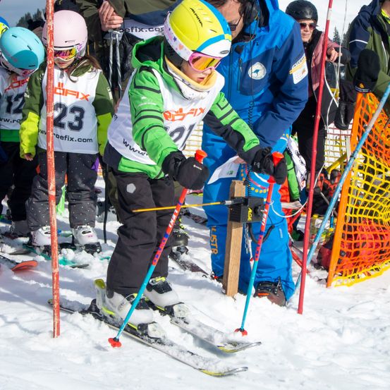 Eine Gruppe von Kindern auf einer Skipiste, die Helme und Skiausrüstung tragen. Sie stehen in einer Schlange und warten auf ihren Zug. Ein Kind hält Skistöcke, während andere umherschauen. Hinter ihnen befindet sich eine Barriere und mehrere Erwachsene in Winterkleidung.