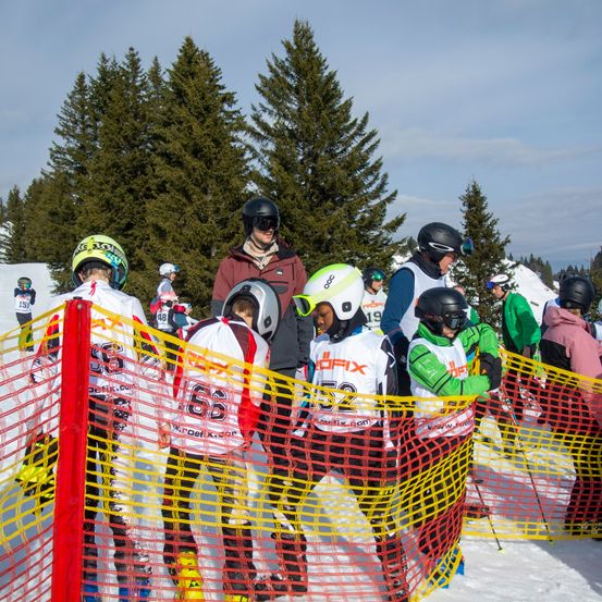 Mehrere Skifahrer in Helmen stehen hinter einer gelben Barriere auf einem verschneiten Hang. Im Hintergrund sind hohe Bäume zu sehen.