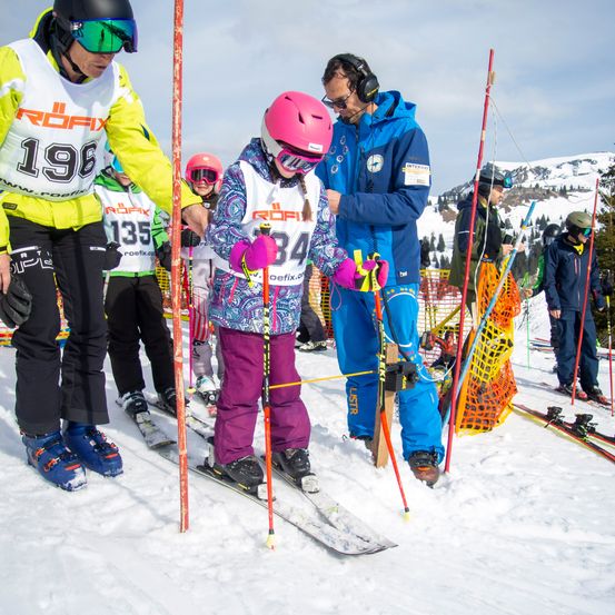 Eine Gruppe von Skifahrern, darunter ein junges Mädchen mit Helm, bereitet sich auf ein Skirennen auf einem verschneiten Berg vor. Ein Lehrer führt sie an.