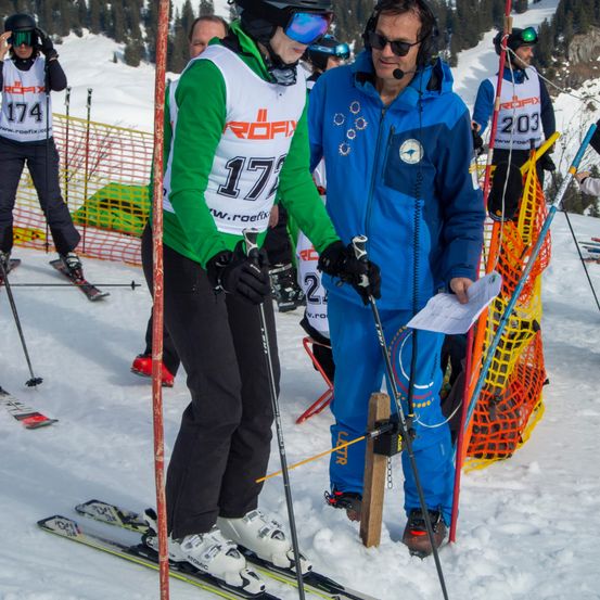 Skifahrer stehen auf einem verschneiten Hang. Ein Skifahrer mit der Nummer 174 hält Skistöcke. Ein anderer Skifahrer mit der Nummer 177 hält ein Papier. Eine Person in einer blauen Jacke hält Skistöcke.