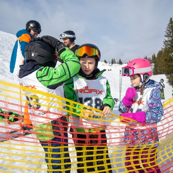 Drei Kinder in Skiausrüstung stehen in einem verschneiten Bereich, tragen Helme und Handschuhe. Der Junge in der Mitte hat eine Brille auf und schaut nach vorne. Die anderen beiden Kinder schauen ihn an. Sie sind von einem gelben Netz umgeben. In der Ferne gibt es Bäume und eine weitere Person.