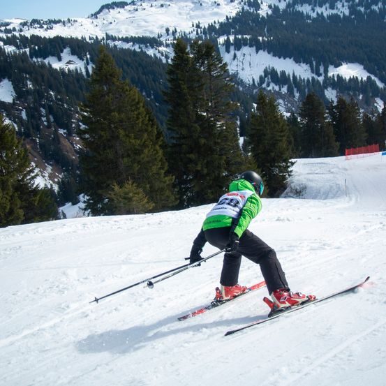 Ein Skifahrer mit einer grün-weißen Jacke mit der Nummer 20 fährt einen schneebedeckten Hang hinunter, mit Bäumen im Hintergrund.