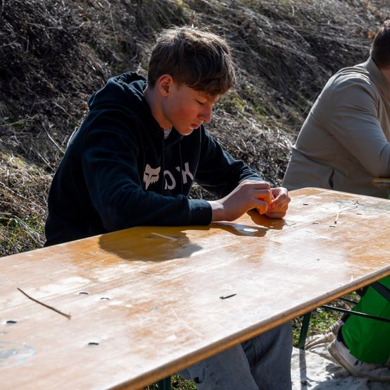 Ein Junge arbeitet an einem Holztisch, während eine weitere Person hinter ihm sitzt. Sie sind draußen, mit getrocknetem Gras und einem Hügel im Hintergrund.