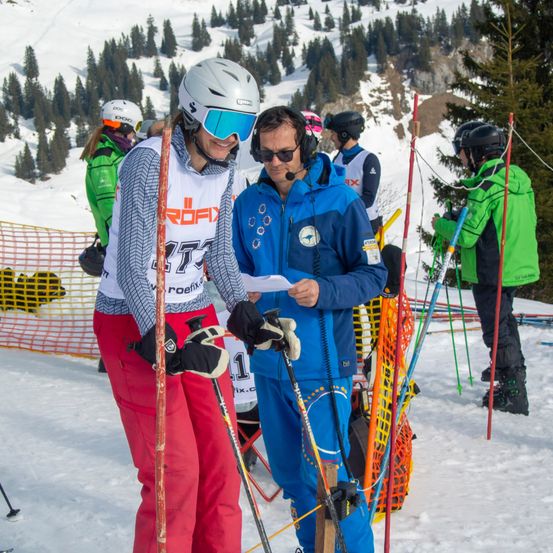 Skifahrer und Snowboarder befinden sich auf einer verschneiten Piste, wobei einige Leute stehen und andere Helme tragen. Eine Person hält ein Papier und scheint zu sprechen.