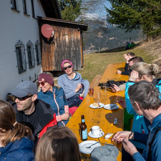 Eine Gruppe von Menschen sitzt draußen an einem Holztisch und lächelt. Hinter ihnen steht eine Frau mit Sonnenbrille und pinker Kopfband. Ein Mann trägt Hut und Sonnenbrille. Eine Flasche steht auf dem Tisch. In der Ferne befindet sich ein Haus mit einer Satellitenschüssel und einer Bergkette.