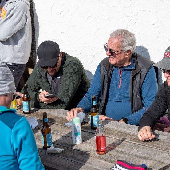 Vier Männer sitzen lächelnd um einen Tisch herum. Zwei haben Bier, einer hält ein Handy, ein anderer eine Saftflasche. Eine weiße Wand ist hinter ihnen.