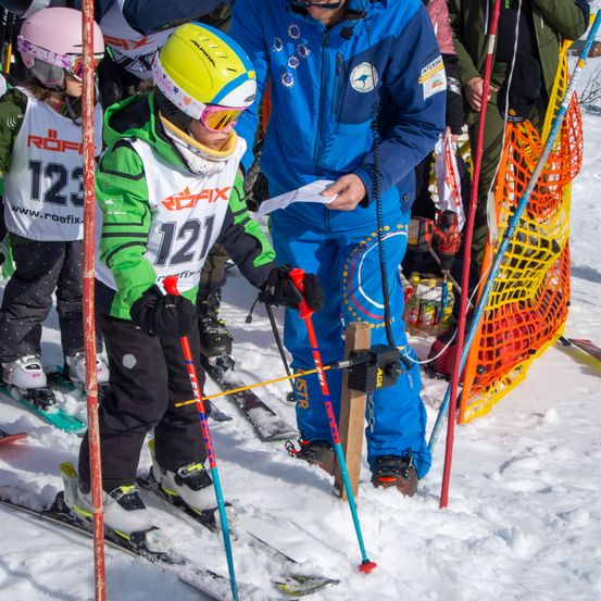 Eine Gruppe von Kindern skifahren auf einem verschneiten Hang und tragen bunte Helme, Skijacken und Schutzbrillen. Ein Kind hält einen Skistock und ein Papier. Ein Erwachsener steht hinter ihnen und hält ein Papier und Skistöcke.