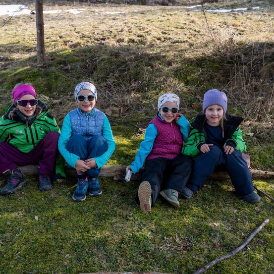 Vier Kinder sitzen auf einem Baumstamm in einem Grasfeld. Sie tragen Winterkleidung und Sonnenbrillen. Sie scheinen für ein Foto zu posieren.
